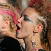 A woman with pink streaks in her hair styled in an updo is singing passionately at Laulupidu. She sports large hoop earrings with multiple dangles and dramatic blue eyeshadow, embodying a Punk vibe. Making a rock on hand gesture, the blurred background suggests an outdoor setting.