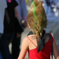 A person with a tall, spiky mohawk dyed with green streaks walks through the crowd at Laulupidu. They are wearing a red tank top and a black bandana with "PUNK" written on it around their neck. The background shows several people dressed casually in various outfits.