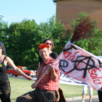 A group of people march outdoors on a sunny day. One person with vibrant red hair and punk-patterned clothing is in focus, holding a flag with anarchist symbols and the words "solidarność". Other participants, dressed in casual attire, walk alongside. Greenery and buildings are visible in the background.