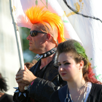 A man with vibrant orange hair styled in a mohawk, wearing sunglasses and a spiked choker, holds a large white flag with colorful designs. Beside him stands a woman with brown hair featuring green highlights, wearing punk-inspired dark clothing and a chain necklace. They appear to be enjoying Laulupidu outdoors.