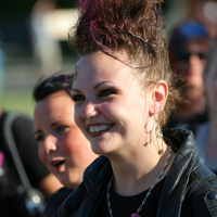 A smiling person with a mohawk dyed purple and black, epitomizing punk style, is wearing a black leather jacket and silver necklace. They have multiple earrings and visible makeup. Another person with a short hairstyle and wearing black can be seen in the background, slightly out of focus, giving the scene a Laulupidu vibe.