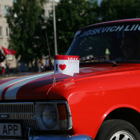 A red vintage car with white racing stripes on the hood is parked. It has several small flags with "I ❤️ TALLINN" and "2011" printed on them, giving it a punk vibe. The front license plate reads "863 APP." The car, part of a gathering akin to Laulupidu, is seen with other cars and people in the background.