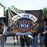 A group of people walking outdoors and holding a banner for "Kära Kants," a hard rock pub established in 2000. The punk-themed banner features flames around a shield-like logo. Participants wear black clothing; one person has a prominent red mohawk, and another has colored, spiked hair. Trees and buildings are visible in the background.