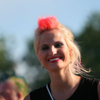 A woman with short, platinum blonde hair featuring a bright pink section at the top is smiling. She is wearing a sleeveless black top with a colorful design on it. The background is an outdoor setting, possibly an Estonian music event, with a clear sky and trees blurred in the distance.