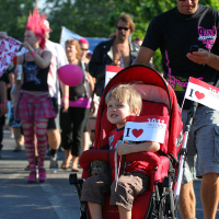 A young child in a red stroller holds a small "I ♥ Tallinn" flag in each hand. The child is pushed by a man wearing sunglasses and a black t-shirt. They are part of the lively Punk Laulupidu street parade, with participants in colorful and eclectic outfits, including pink plaid skirts and flashy accessories.