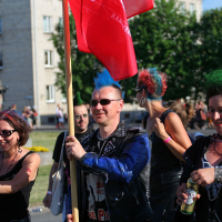 A group of people with colorful mohawk hairstyles and punk outfits are walking outdoors, holding a red flag. They seem to be part of a Punk Laulupidu, with onlookers and buildings visible in the background. Some are smiling and appear to be in high spirits.