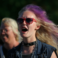 A person with long, wavy, blonde hair partially dyed purple is wearing red-framed sunglasses and a black choker with a circular ring. They appear to be mid-speech or singing with their mouth open, evoking the spirit of Laulupidu. Dressed in a denim vest, they stand outdoors with another individual blurred in the background.