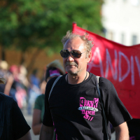 A group of people walk in a street parade. The man in the center wears sunglasses, a black t-shirt with colorful text, and carries a backpack. A woman next to him, with a punk vibe, wears a bandana and necklace. In the background, another person holds a red banner that partially reads "DIVER." Trees are in the distance.