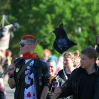A group of people, some with brightly colored hair and black leather clothing, are marching outdoors. One person with short red hair carries a black flag with a skull design, evoking a punk Laulupidu vibe. Another person carries an animal skull on a stick. Trees and a building are visible in the background.