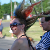 A punk with colorful dyed hair and sunglasses smiles while standing outdoors. Their hair is styled in spikes with red, blue, and green colors. They wear large earrings and a halter top. Another person in a black and white striped shirt stands next to them. Trees and other people are visible in the background, giving a festive Laulupidu vibe.