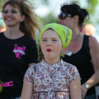 A young girl wearing a floral dress and a yellow bandana stands outdoors with her mouth slightly open, bringing an unexpected punk flair to the scene. Behind her, several adults and another child are visible, some smiling. The background is slightly blurred, suggesting movement and a lively Laulupidu atmosphere under bright sunlight.