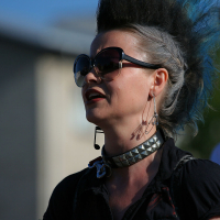 A person with a tall black and grey mohawk hairstyle is seen outdoors, embodying a punk aesthetic. They wear large, dark sunglasses, long earrings, and a studded choker necklace. The background shows blurred buildings and greenery, with another person partially visible behind them. The lighting suggests it is daytime.