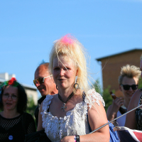A group of people with colorful, punk-inspired hair, some sporting spiked and dyed styles, are enjoying an outdoor event on a sunny day. A woman in the foreground wears a white lace top, with pink and blue streaks in her hair. The lively scene resembles the festive spirit of Laulupidu against a backdrop of buildings and blue sky.