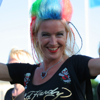 A person with brightly colored, rainbow hair smiles widely. They are wearing a punk-inspired black Ed Hardy shirt with skull designs and a heart-shaped necklace, raising one arm enthusiastically. The background is slightly blurred with blue and white Laulupidu flags and another person partially visible.