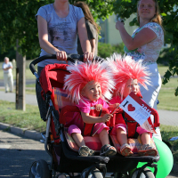 A smiling woman in a punk-inspired outfit is pushing a double stroller with two young children. The children are wearing vibrant red costumes with large, red-and-white striped wigs and holding a sign that reads "I ♥ DAD." Another woman, smiling, stands nearby. They are outdoors on a sunny day with trees in the background.