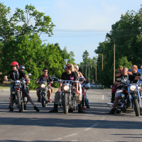 A group of motorcyclists, including one with a red helmet, are lined up on a road in front of a multi-story building and surrounded by tall trees. There are six motorcycles with riders sitting on them, reminiscent of a punk gathering, while people casually gather around or walk in the background.