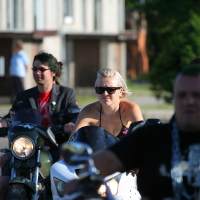 A group of motorcyclists rides down a street on a sunny day. At the front, a punk woman in sunglasses and a black sleeveless top rides a white motorcycle. A man next to her wears a dark jacket and sunglasses on a black motorcycle. Green trees and buildings are in the background, contrasting with their edgy style.
