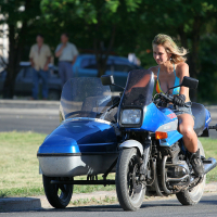 A woman with blonde hair, wearing a punk-inspired blue top and black shorts, rides a punk-style blue motorcycle with a sidecar on a paved road. The background is blurred with trees and two pedestrians, suggesting motion. The scene feels energetic as if capturing the spirit of Laulupidu during daylight.