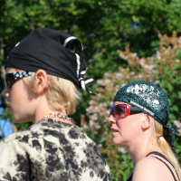 Two people outdoors on a sunny day, both wearing black bandanas and sunglasses. One sports a black and white tie-dye shirt with a chunky necklace, while the other rocks a punk-inspired black tank top. Lush green foliage surrounds them, reminiscent of Laulupidu's natural beauty. They seem focused on something off-camera.