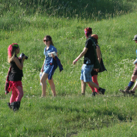 A group of people, five adults, are walking through a grassy field on a sunny day. They are dressed in a mix of casual and colorful attire, with some wearing hats and bright leggings reminiscent of Punk Laulupidu festivity. One person is holding a camera and appears to be taking photos. The field is lush and green.