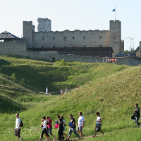 A group of people walks along a path through a grassy hillside toward a large, medieval stone castle with multiple towers, reminiscent of scenes from Laulupidu celebrations. Some people are visible on a higher path closer to the castle. One tower has a flag at the top, and the sky is clear and blue in the background.