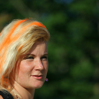 A woman with blonde hair featuring bright orange streaks stands outdoors, exuding a punk vibe. She is wearing a black top and has a small pink earbud in her left ear. The blurred background of green foliage and sunlight adds to the Laulupidu-like atmosphere. She has light skin, freckles, and an expression suggesting she is speaking or listening.