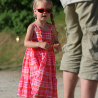 A young girl wearing a pink plaid dress, pink sandals, and red sunglasses stands outdoors holding colorful beads during Laulupidu. An adult, partially visible from behind, stands nearby in beige pants and sandals. The background features greenery with sunlight filtering through.