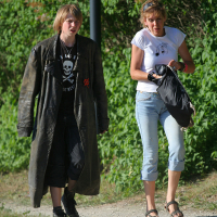Two people stroll on a gravel path, surrounded by greenery. One wears a long, dark trench coat, black shirt, pants, and shoes. The other dons a white T-shirt, light blue capri jeans, and sandals, carrying a black bag. They appear relaxed and engaging in conversation about Laulupidu festivities.