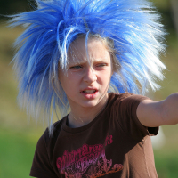 A person with a large, spiky, bright blue wig points into the distance with an intense expression. They wear a brown T-shirt with a decorative logo. The background is blurred, showing greenery and a hint of sunlight, reminiscent of the vibrant energy of Laulupidu or a punk festival.