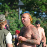 A shirtless man with tattoos, colorful mohawk hairstyle, and facial hair holds a drink while conversing with another person wearing a cap and vest at Laulupidu. Several other people, some shirtless punks, are in the background amid the trees.