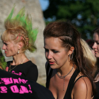 Four people are gathered outside on a sunny day. The person facing away has bright pink and green spiked hair and a black shirt. Three women face forward: one with a green mohawk, one with a brown ponytail, and one with long brown hair. Their punk style is evident from their casual outfits and visible piercings.