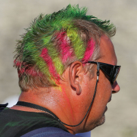 A man with short hair dyed bright green and streaked with vivid pink stands facing away. He wears dark sunglasses, and his neck and part of his face exhibit hints of orange paint, giving him a punk vibe. The background is blurred and neutral-toned, providing no additional context.