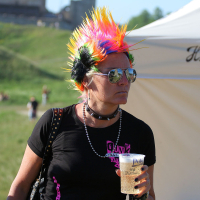 A punk woman sporting a vibrant rainbow mohawk, sunglasses, and a black choker is holding a plastic cup of beer. She is wearing a black T-shirt with pink and white text. In the background, at Laulupidu, there is a white tent with writing and a grassy area with people in the distance.