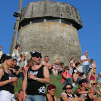 A group of people, many wearing matching black and white tank tops and baseball caps, sit and stand on a grassy hill, enjoying drinks. Behind them is a stone tower structure with a wooden roof against a clear blue sky. Some have sunglasses, others hold cups while conversing in a laid-back punk vibe at Laulupidu.