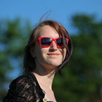 A person with shoulder-length hair wearing bright red sunglasses is smiling slightly while standing outdoors. Dressed in a black lace top with gold buttons, they exude a punk vibe. The background features blurred, green, leafy trees against a clear blue sky, reminiscent of a vibrant Laulupidu scene.