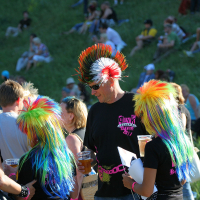 People at an outdoor event, some sporting colorful rainbow mohawk wigs and black shirts with pink text, evoke a punk spirit. One individual, adorned with a black mohawk and sunglasses, stands out holding drinks. In the background, groups of people are sitting and relaxing on a grassy hill at Laulupidu.