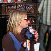 A woman with blonde hair and sunglasses, wearing a white sleeveless shirt, is holding a package of "PUNK" coffee beans and smiling. She stands next to a stack of brown plastic crates filled with additional packages of the same coffee. The background features blurred people and green grass, evoking the spirit of Laulupidu.