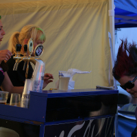 A punk woman with a green and red mohawk bends down dramatically near a bar setup, while two women in black shirts stand behind the counter, one operating beer taps. Another person watches on from the right, under a blue tent. The scene feels reminiscent of Laulupidu, set at an outdoor event or festival.