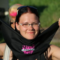 A smiling woman with glasses and dark shoulder-length hair holds a black bandana with pink writing under her chin. She is outdoors in a sunny environment, possibly at a Laulupidu festival, with greenery in the background. She wears a punk-inspired silver watch on her left wrist and has her nails painted a light color.