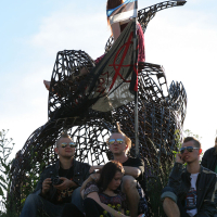 A group of young people, embracing a punk aesthetic, lounges around a large, abstract metal sculpture in a grassy area. One person is perched atop the structure, holding a flag that echoes the Estonian Laulupidu spirit with an X on a white field and bottom resembling the Estonian flag. The sky is clear and bright.