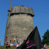 People are gathered on a grassy hill in front of an old, weathered windmill with a wooden structure at the top. A black flag with pink and white text is prominently waving in the foreground, lending a punk vibe to the scene. The sky is clear and blue, indicating a sunny day reminiscent of Laulupidu celebrations.
