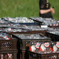 Plastic crates filled with clear plastic bags, each containing white tubes labeled "Пломбир Пломбир" (Plombir), are arrayed in a field under sunlight. Amid the setting reminiscent of a Laulupidu celebration, a person in a dark outfit stands on green grass, blurred in the background.