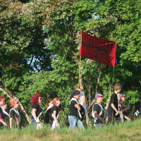 A group of people march outdoors on a grassy terrain, reminiscent of the spirit at Laulupidu. They are holding a red banner with the words "In ternat's krisehnsis" written in black. Dressed casually in primarily black t-shirts and jeans, they walk among green trees with white blossoms, suggesting a spring or summer setting.