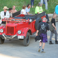A group of people gathers around a vintage red open-top vehicle, which has a spare tire mounted on the side. Some are seated inside while others stand nearby on a concrete surface, embodying a laid-back punk vibe. The backdrop features greenery and a stone wall, with more individuals sitting on the grass in the background, reminiscent of Laulupidu's communal spirit.