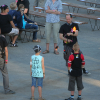 A person dressed in black is juggling flaming torches while three young people with a punk style watch, standing in a circle around the performer. In the background, other people are sitting or standing near wooden picnic tables, engaging in conversation or observing the performance.