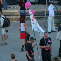 A group of people at an outdoor event blends punk and tradition. One person with a red mohawk and red skirt walks away holding pink balloons. Another person in front holds a large white flag with writing. Several individuals wear black shirts with bright pink text. Stage equipment and speakers are visible in the background, reminiscent of Laulupidu's vibrant atmosphere.