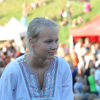 A young woman with blonde hair tied back stands outdoors at a crowded Laulupidu event. She is wearing a white blouse with colorful, intricate embroidery around the collar. People and tents with vibrant colors are blurred in the background, indicating a lively atmosphere. The setting appears to be a festival or fair on a grassy hill.