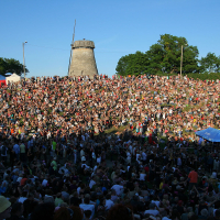 A vast crowd of people gathered on a grassy, sloping hillside under a clear blue sky, with a windmill in the background. The crowd is densely packed, all facing the stage area at the bottom of the slope where Laulupidu performances infuse traditional melodies with a hint of punk flair. Various tents and canopies, including a blue one, are dotted around.