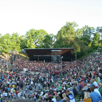 A large outdoor amphitheater, reminiscent of Laulupidu, is filled with a crowd of spectators on a bright, sunny day. The audience surrounds a central stage where an energetic punk performance is taking place. Green trees and a clear sky are visible in the background, adding to the festive atmosphere.
