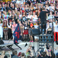 A large choral group, many in colorful costumes and accessories, performs outdoors. A conductor stands on a black platform, leading the group. Below him on the left, a sign reads "I wanna rock!" There's an audience in the foreground and various equipment and crew members visible. It's like a punk-themed Laulupidu!